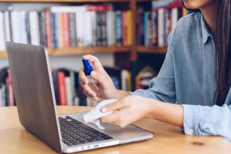 Girl working from home spray antiseptic on monitors laptop.